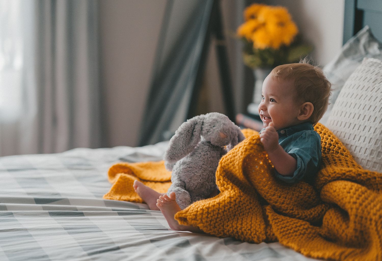 Laughing baby wrapped in yellow knit blanket cuddling stuffed elephant toy on cozy bed.