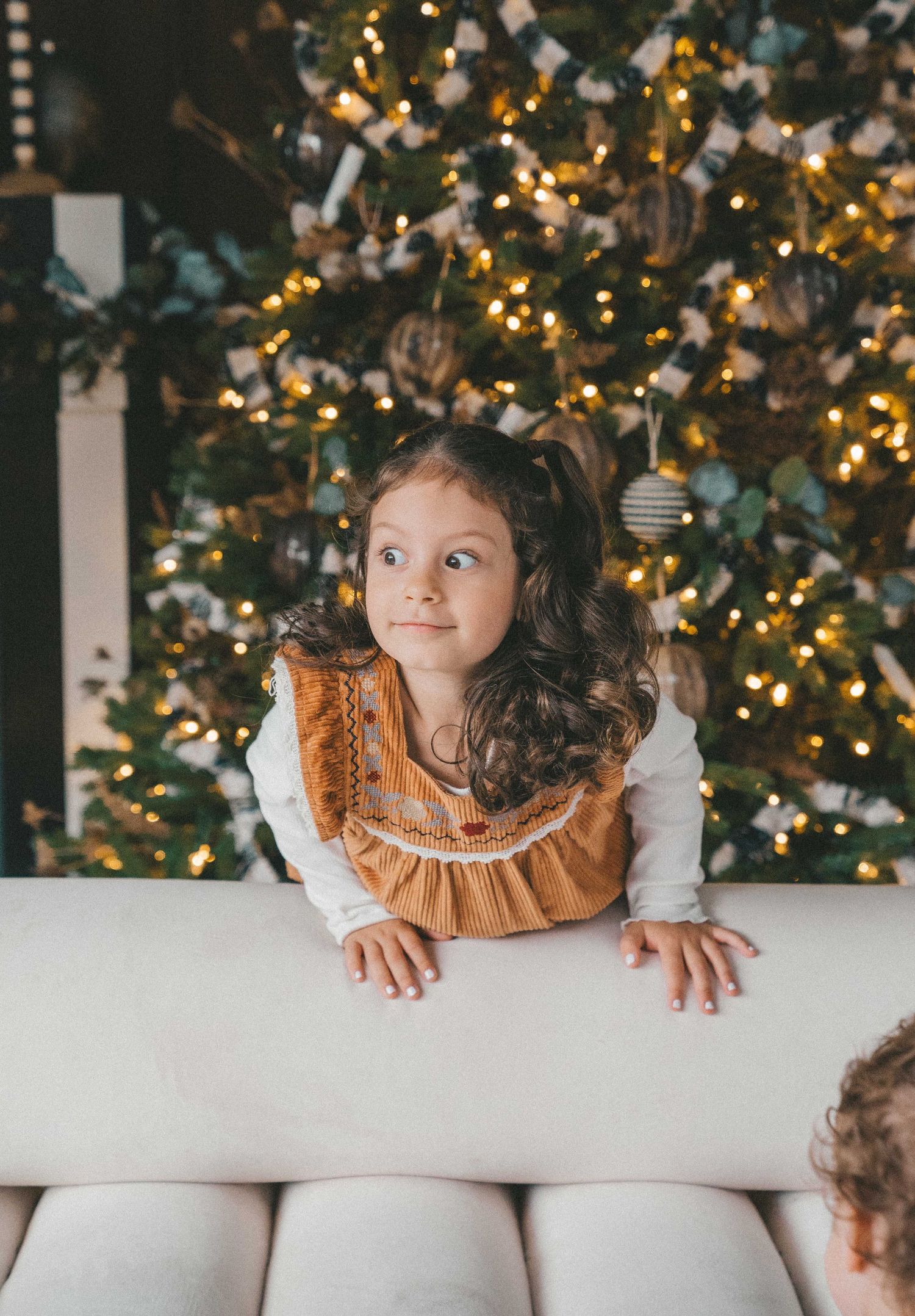 A little girl in a brown dress stands by a glowing decorated Christmas tree, looking up curiously.