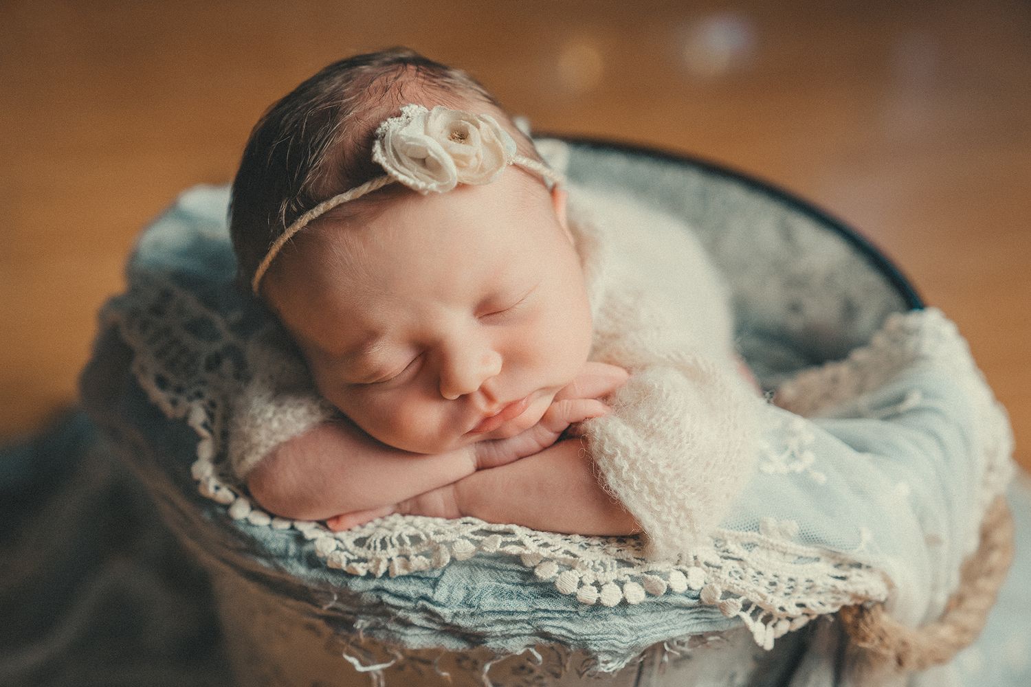 Sleeping newborn girl with a floral headband nestled in a rustic bucket with lace wrap.
