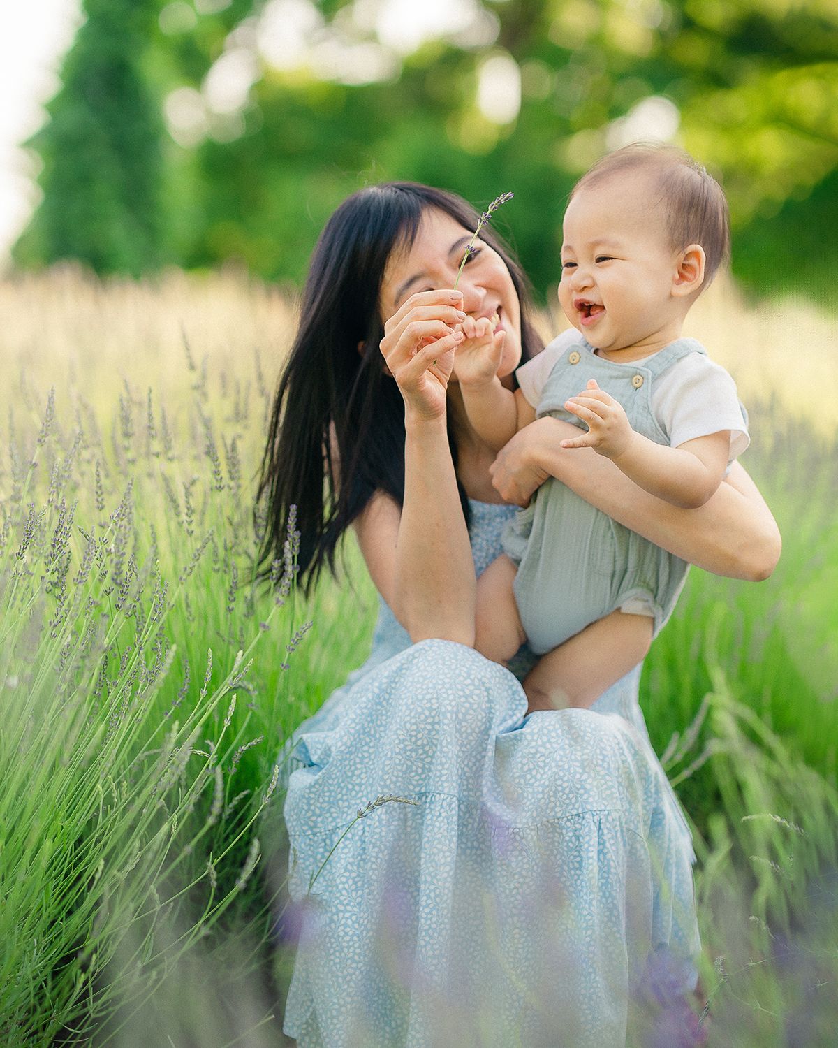 A smiling mother in a blue dress holds her laughing baby in a fragrant lavender field.