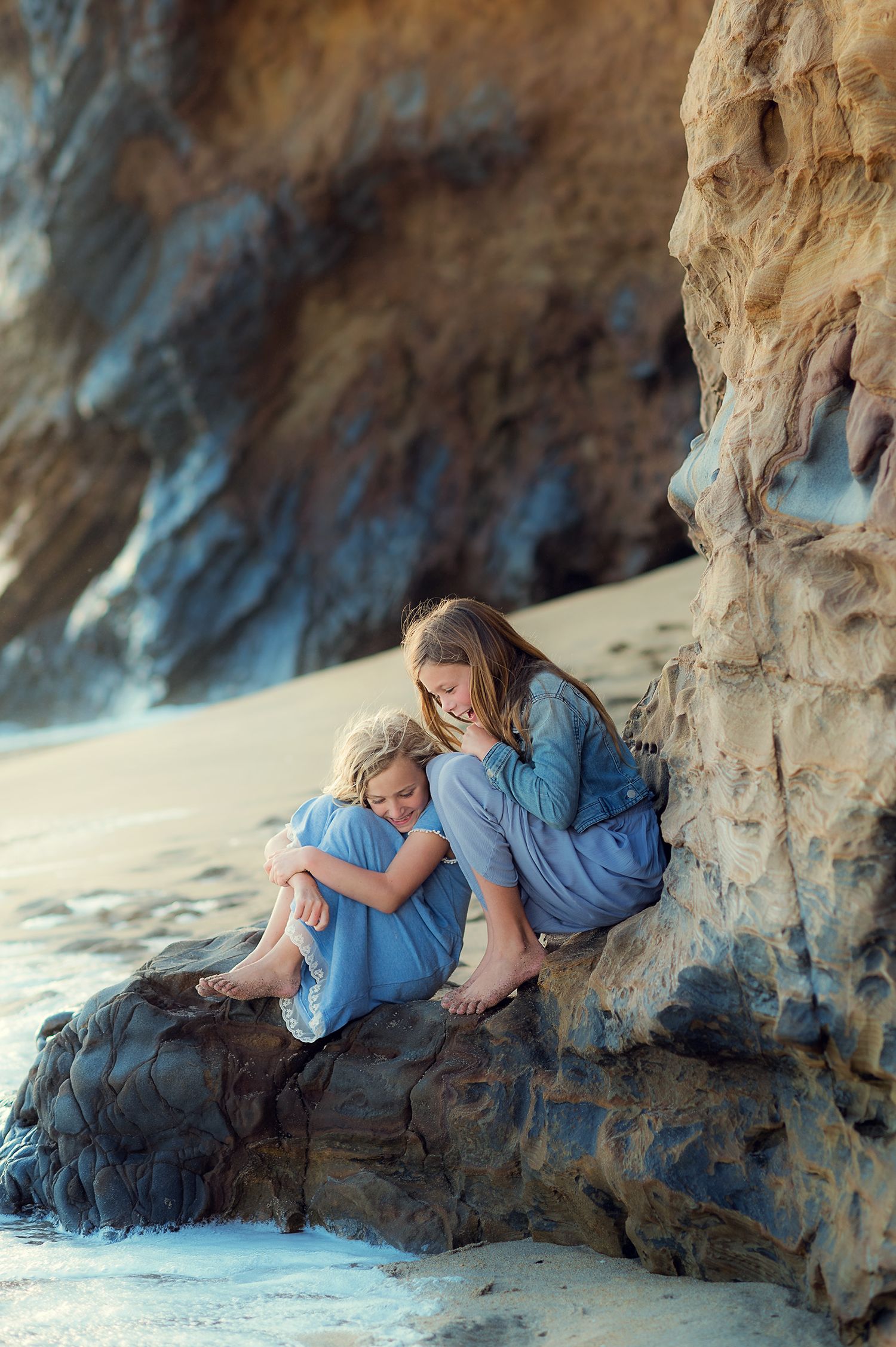 Two girls in blue dresses share a tender embrace while sitting on rocks at a scenic beach.