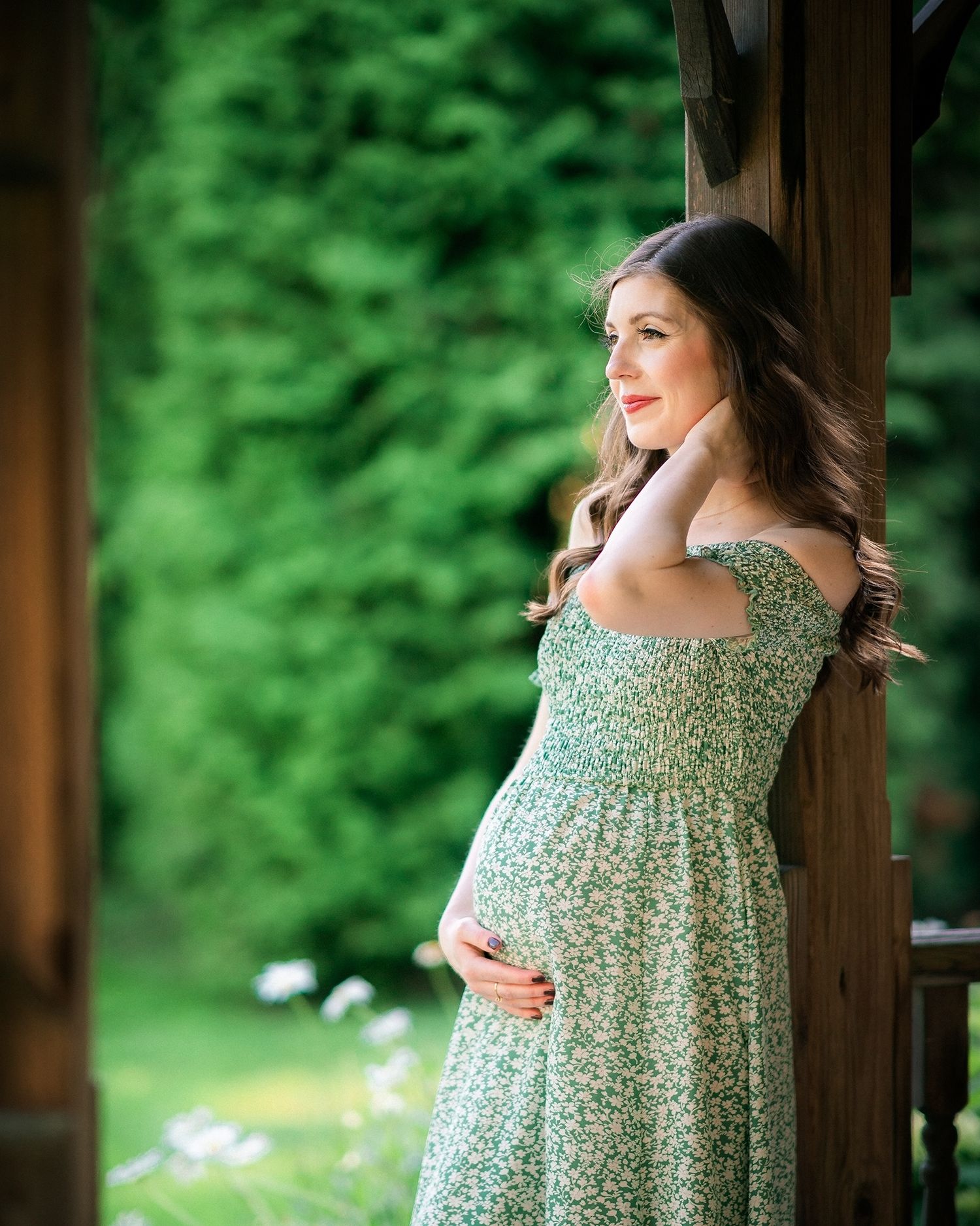 Pregnant woman in green floral dress holding belly while leaning against wooden post.