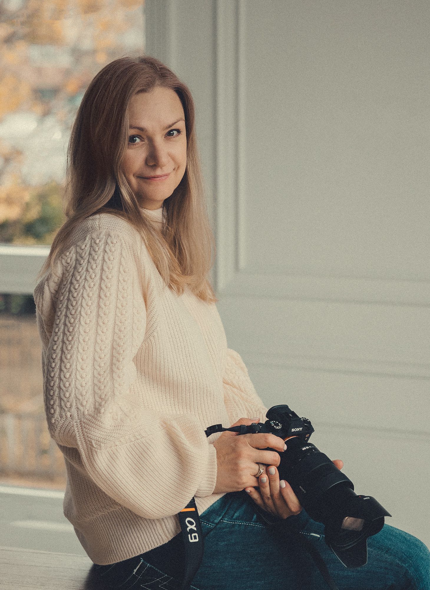 Smiling female photographer holding a camera while sitting by a bright window indoors.