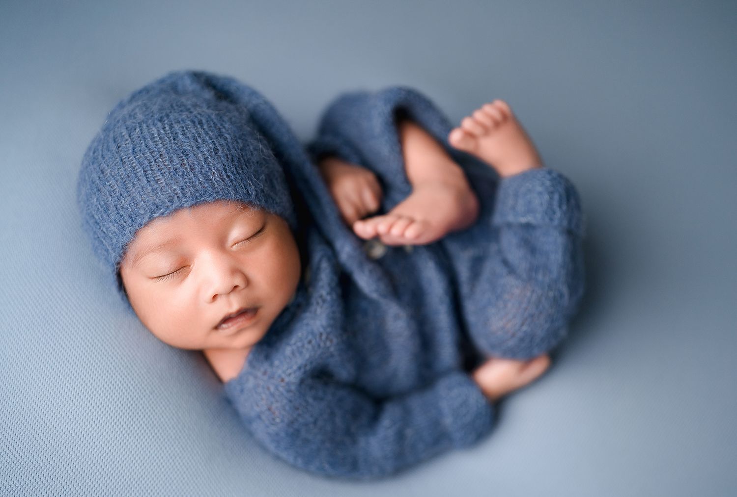 Sleeping newborn baby in a blue knit hat and matching romper curled up in a cozy pose on a soft blue backdrop.