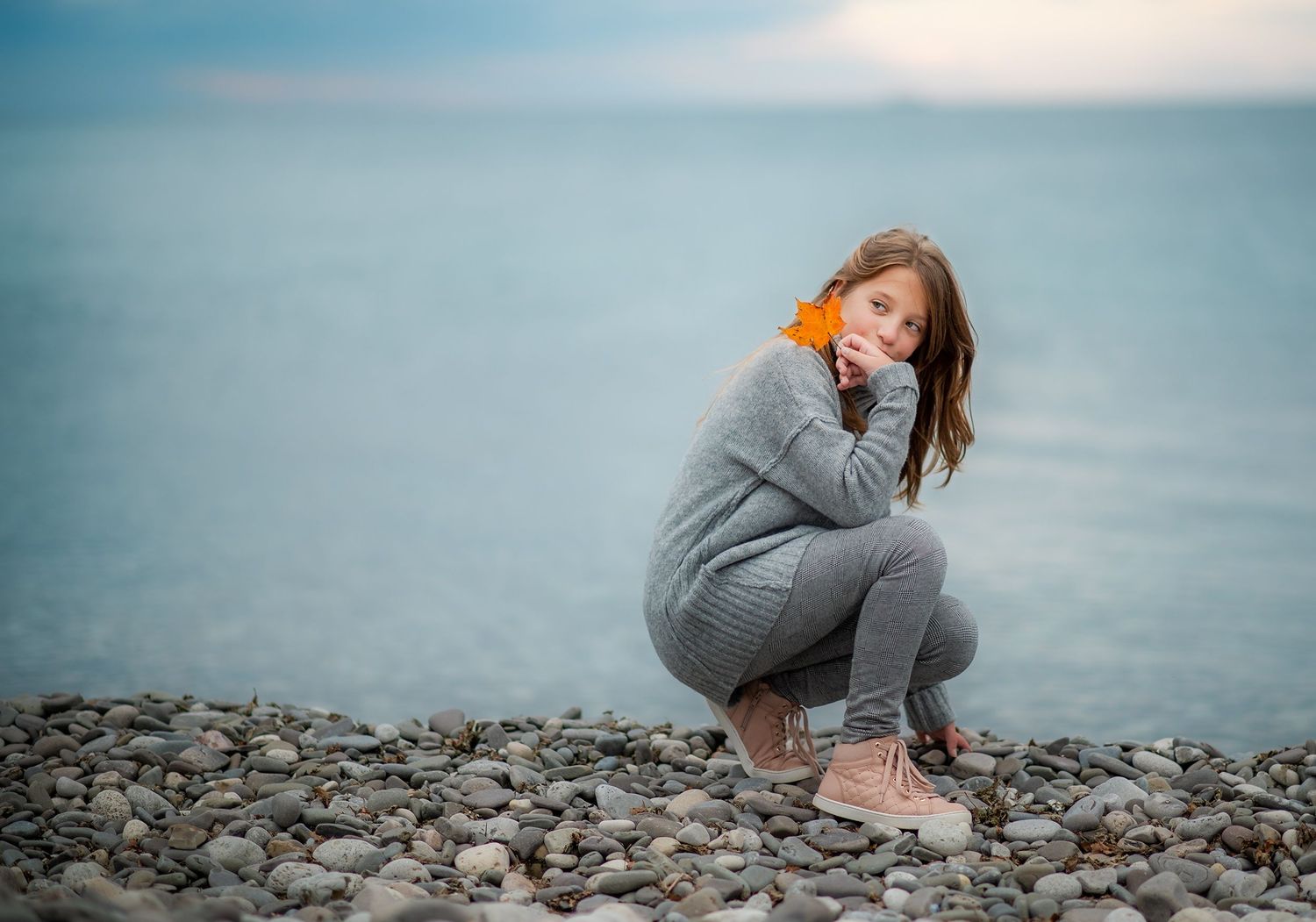 Young girl in gray outfit crouching on rocky pebble beach beside calm blue water.