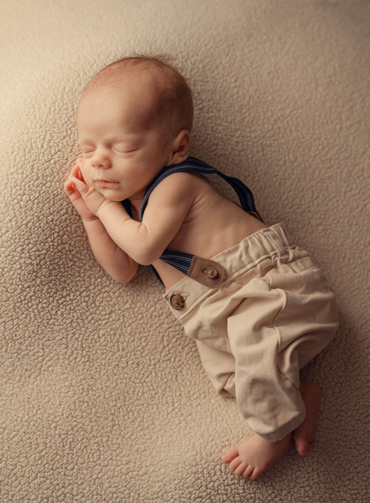 Newborn baby boy sleeping peacefully wearing suspenders and khaki pants on a cream blanket.