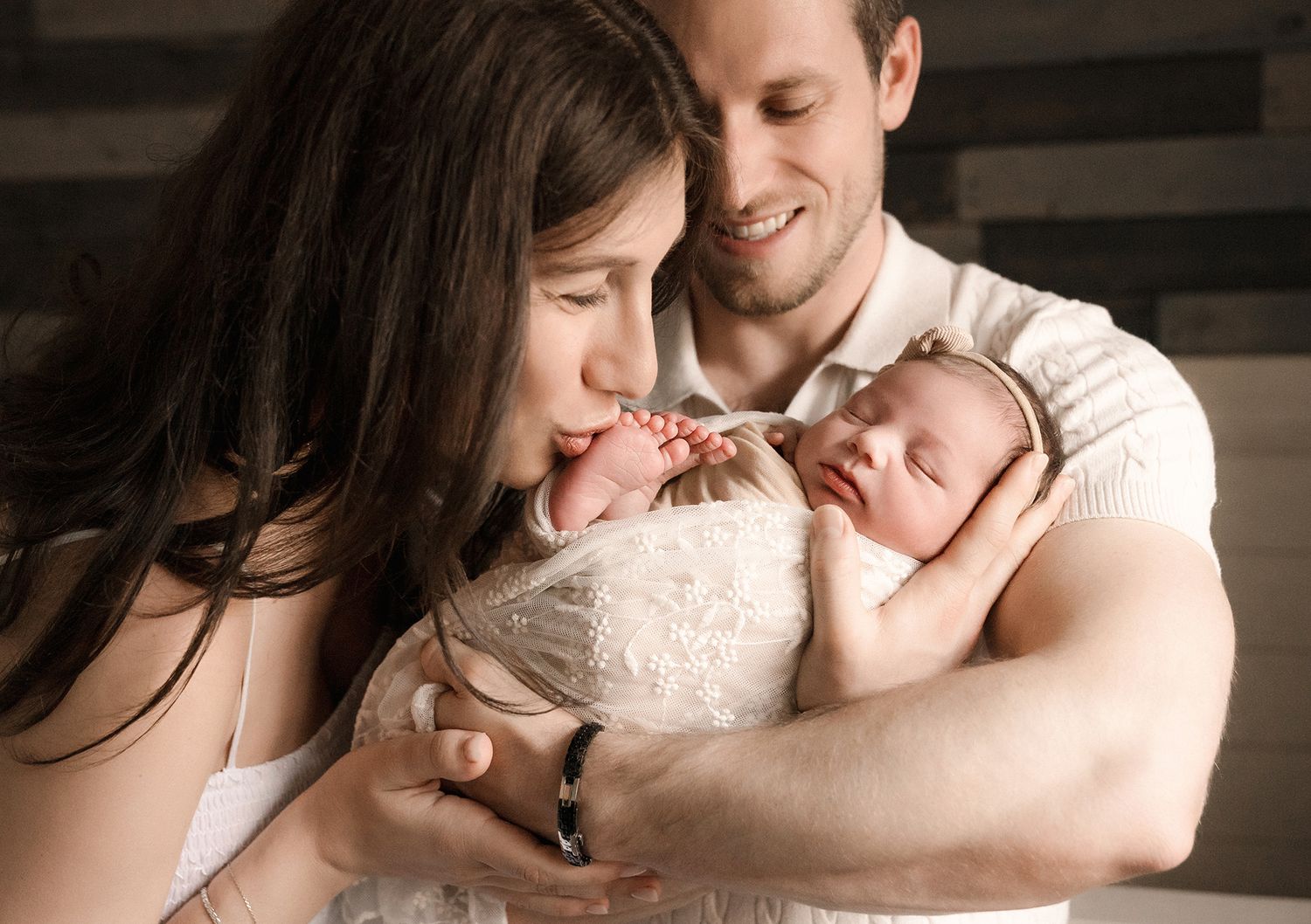 Smiling parents holding swaddled newborn as mother gives baby a gentle kiss.