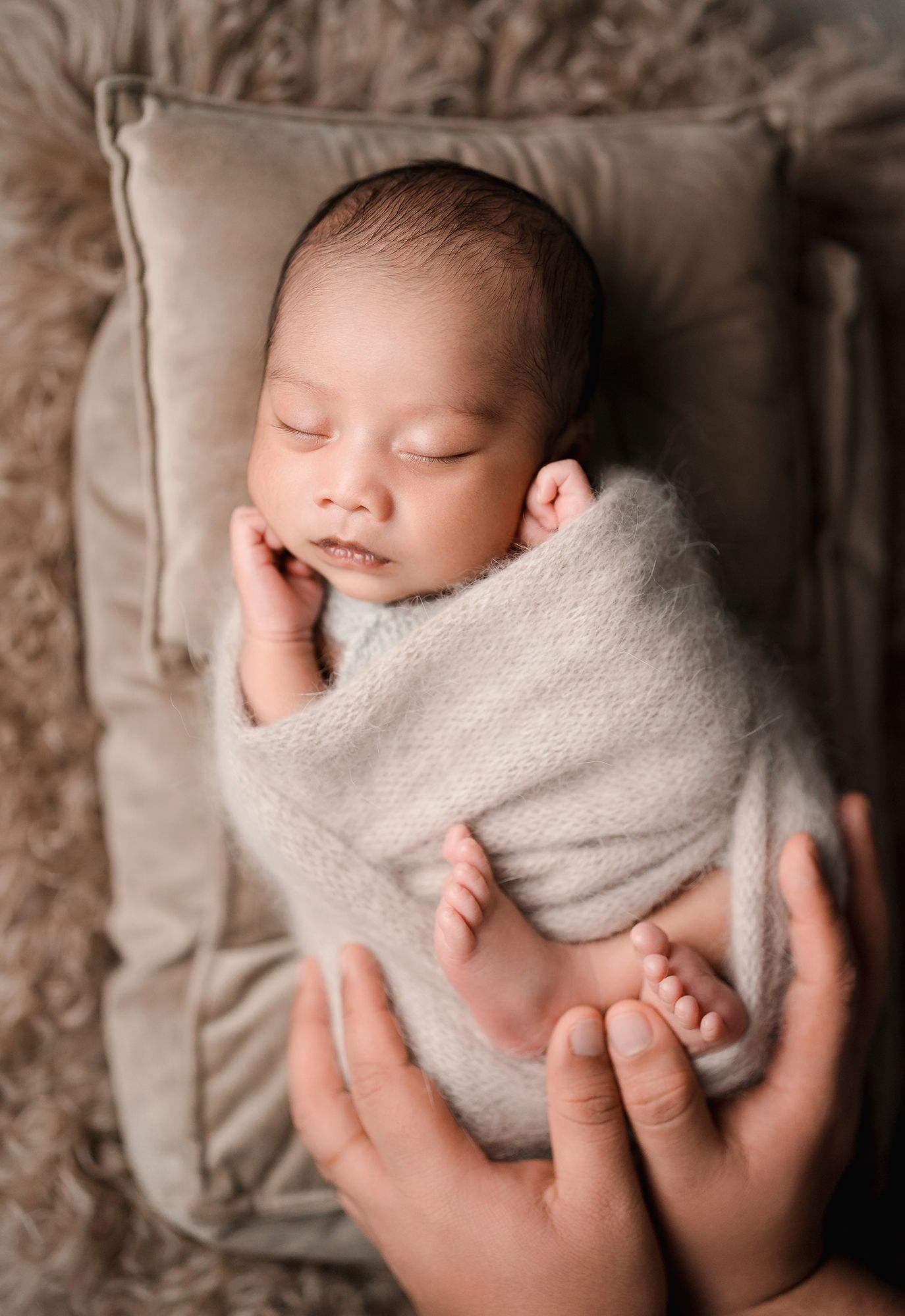Sleeping newborn baby wrapped in a soft gray knit swaddle resting on a cushion while parents' hands gently support from below.