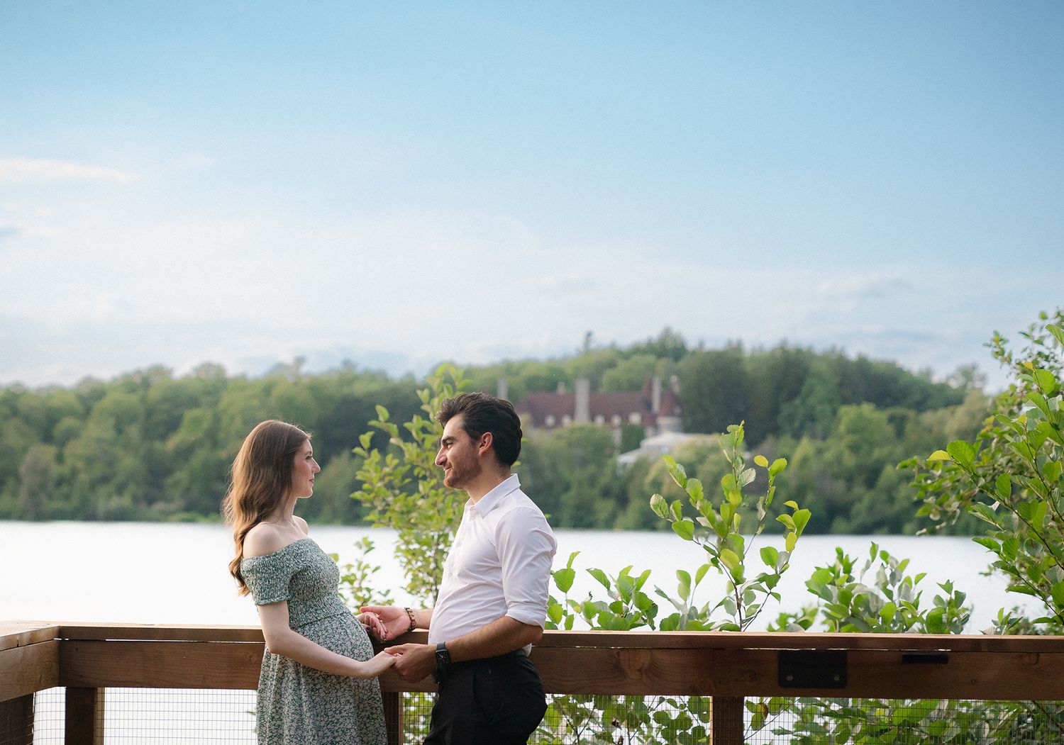 Expecting couple holding hands on wooden deck overlooking scenic lake.