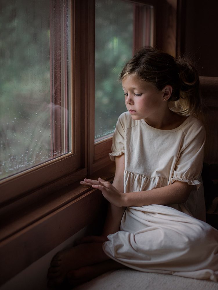 A young girl in a white dress sits by a rain-speckled window, gazing outside thoughtfully.