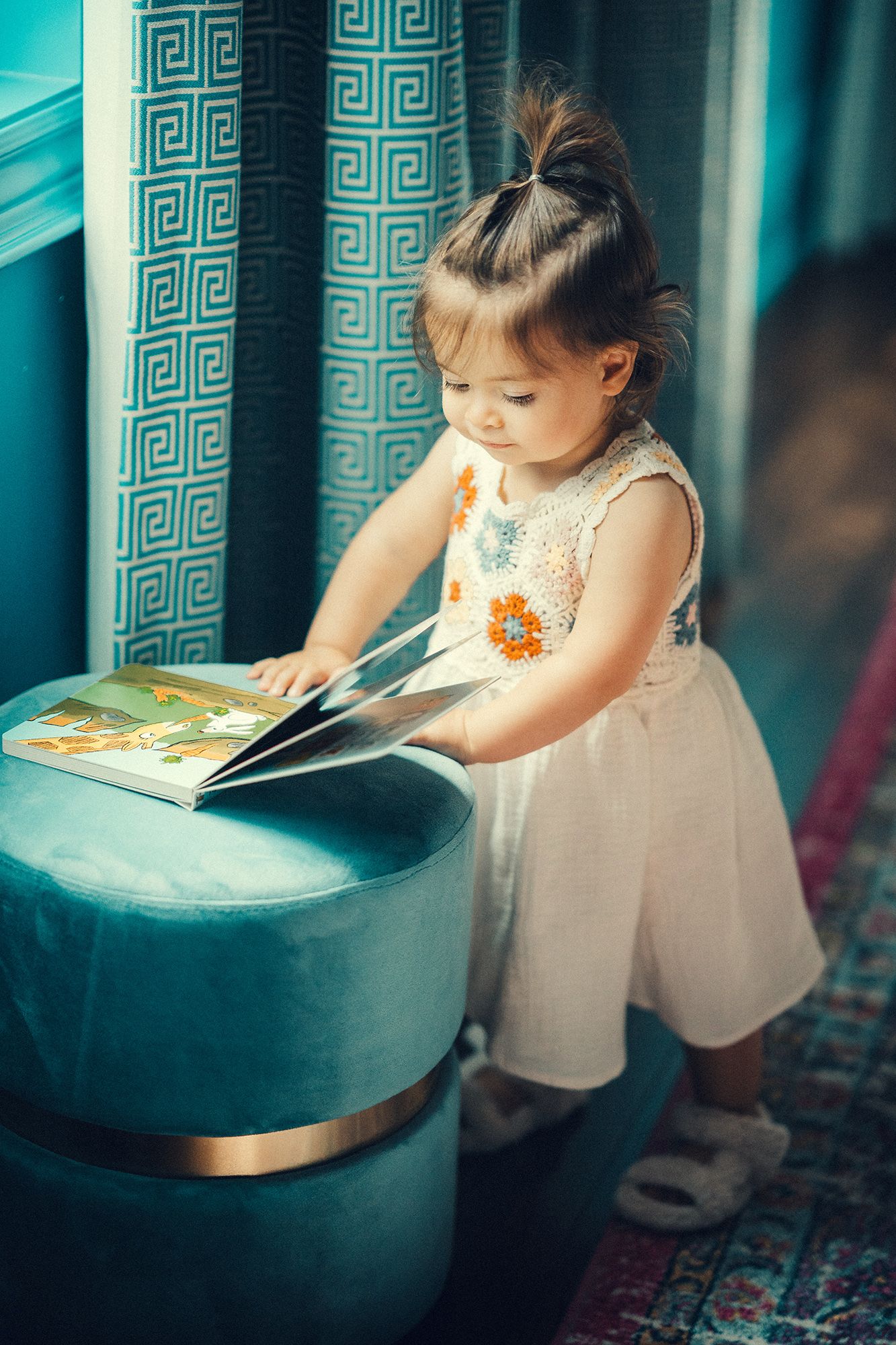 A little girl in a white dress carefully flips through a colorful picture book on a teal ottoman.