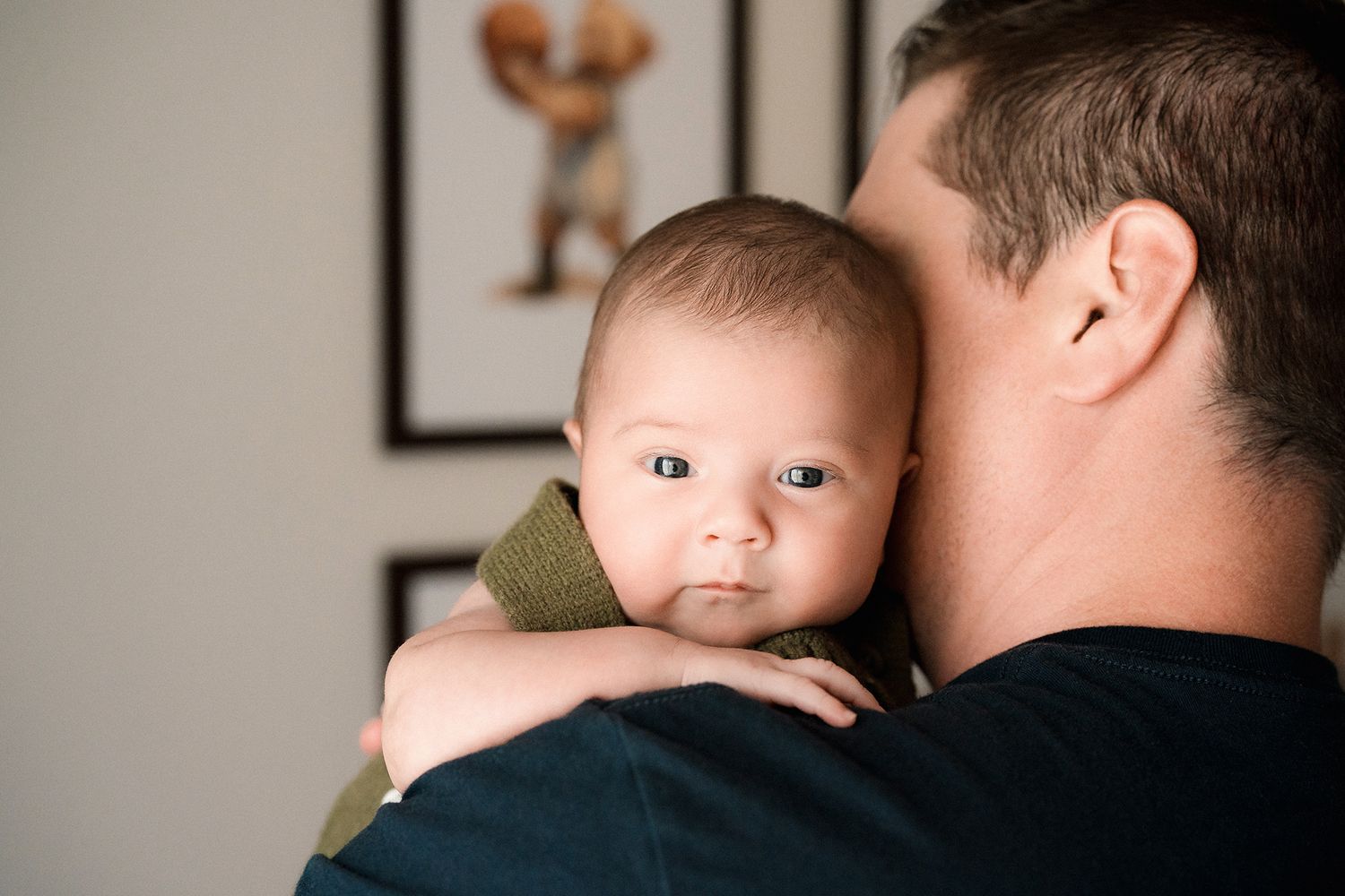 Wide-eyed baby in an olive green outfit rests against the shoulder of a dark-haired man in a navy shirt, with framed art behind them.