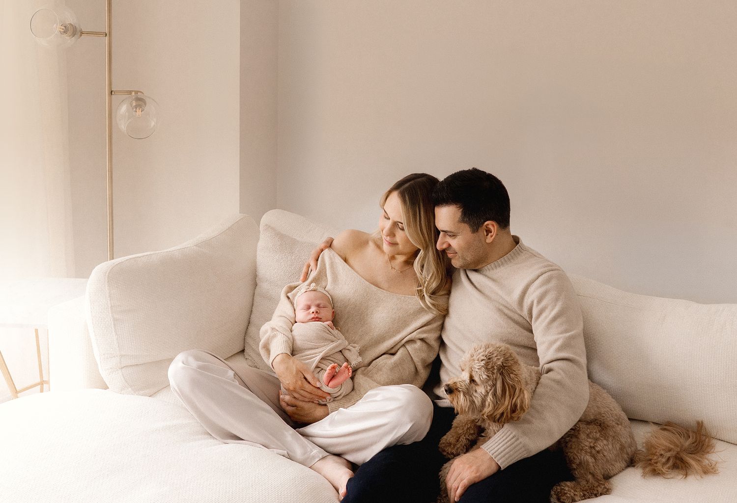 A smiling couple cuddles their newborn baby and golden dog on a white sofa in a bright space.