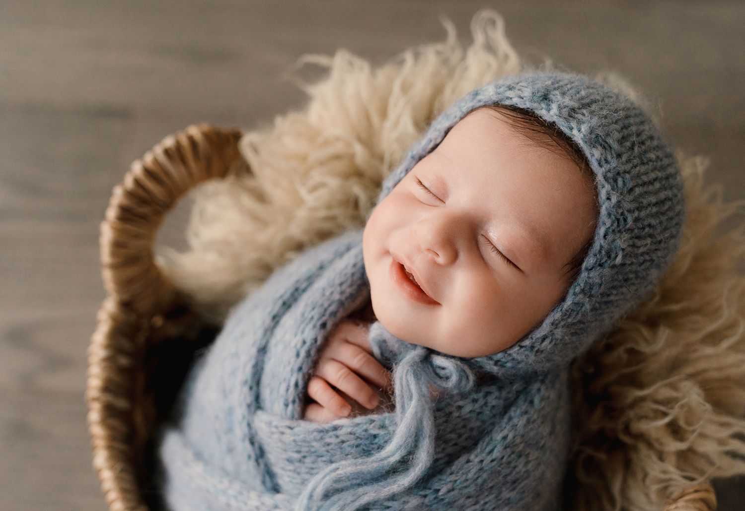 Smiling newborn baby wrapped in blue knit swaddle and bonnet in a wicker basket.