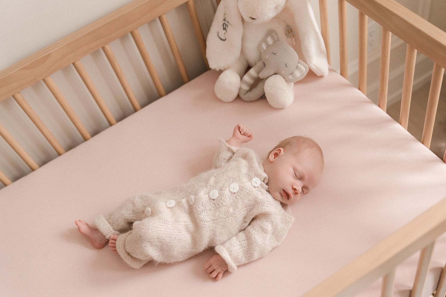 Sleeping baby in a cream knit romper lying on a pink sheet in a wooden crib with plush bunny toys.