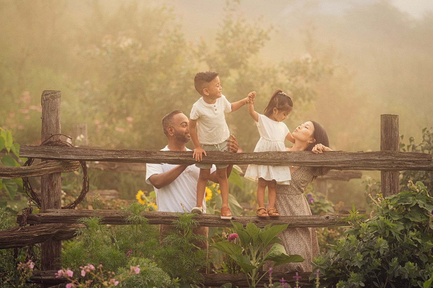Joyful family of four playing on a rustic wooden fence in a golden misty garden.