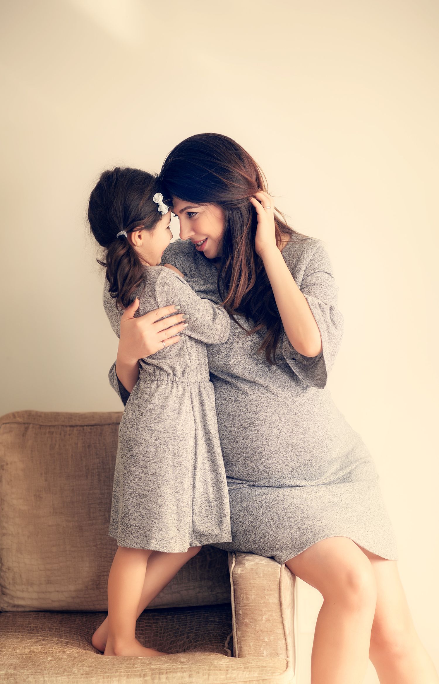 Pregnant mother in gray dress hugging young daughter on couch in warm indoor setting.