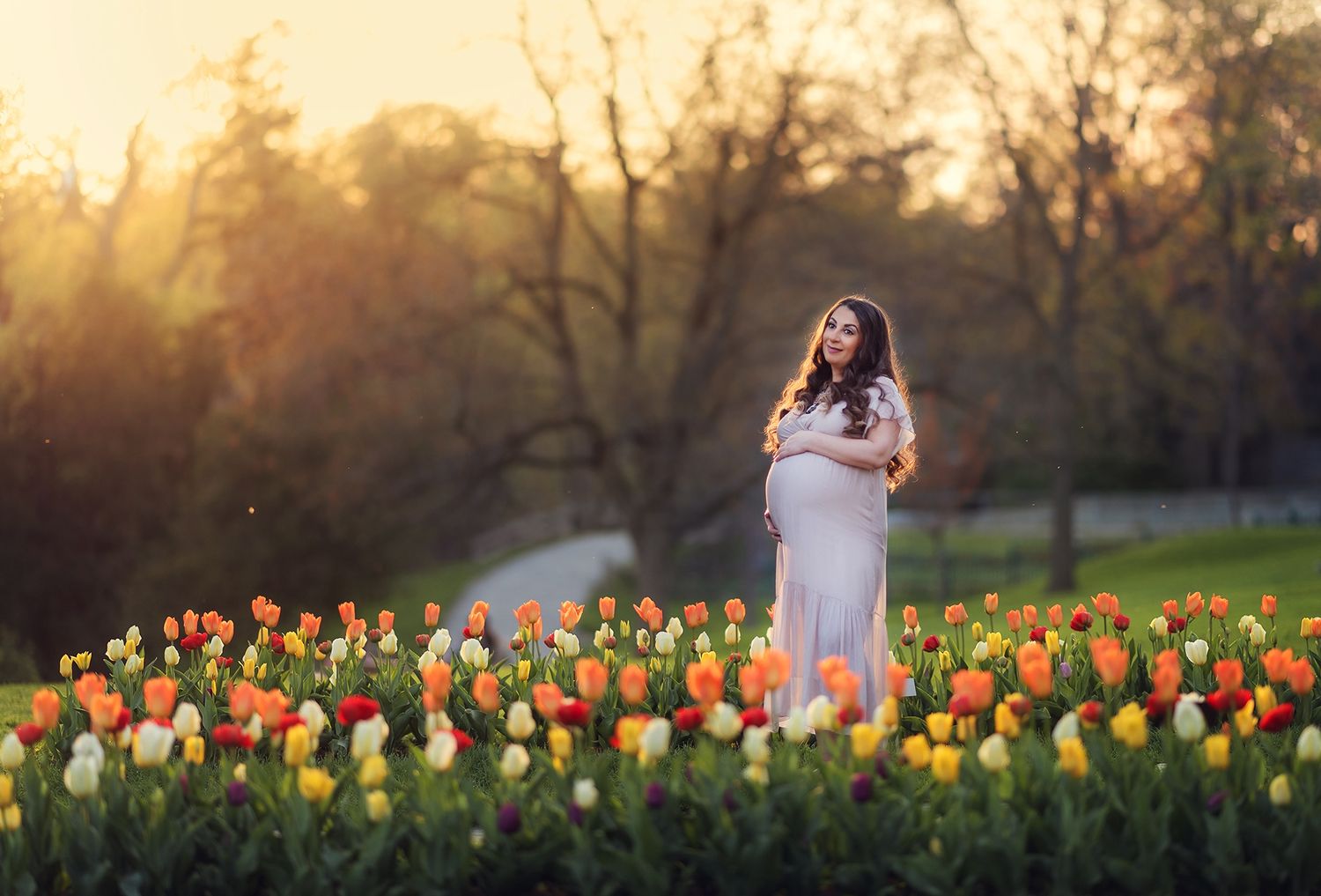 Pregnant woman in white dress standing in colorful tulip garden at golden hour.
