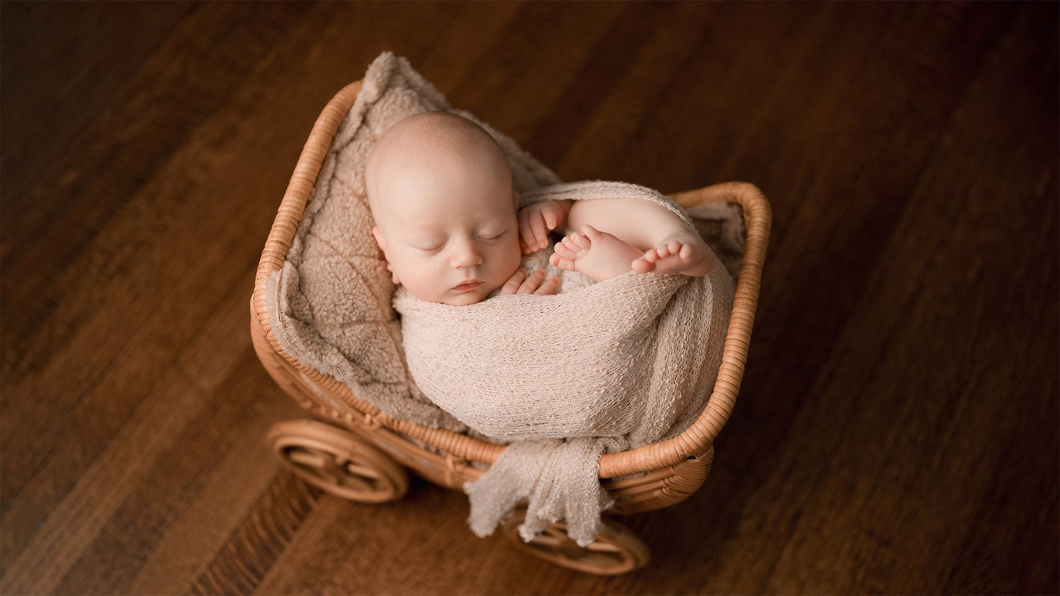 Sleeping newborn baby wrapped in beige swaddle resting in a wicker pram prop on wood floor.