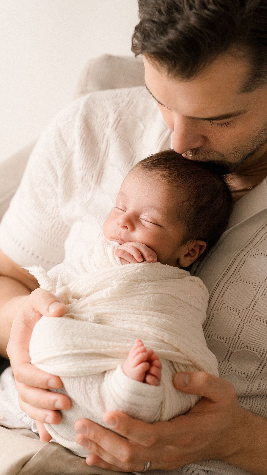 Father tenderly kisses sleeping newborn baby wrapped in white swaddle blanket.