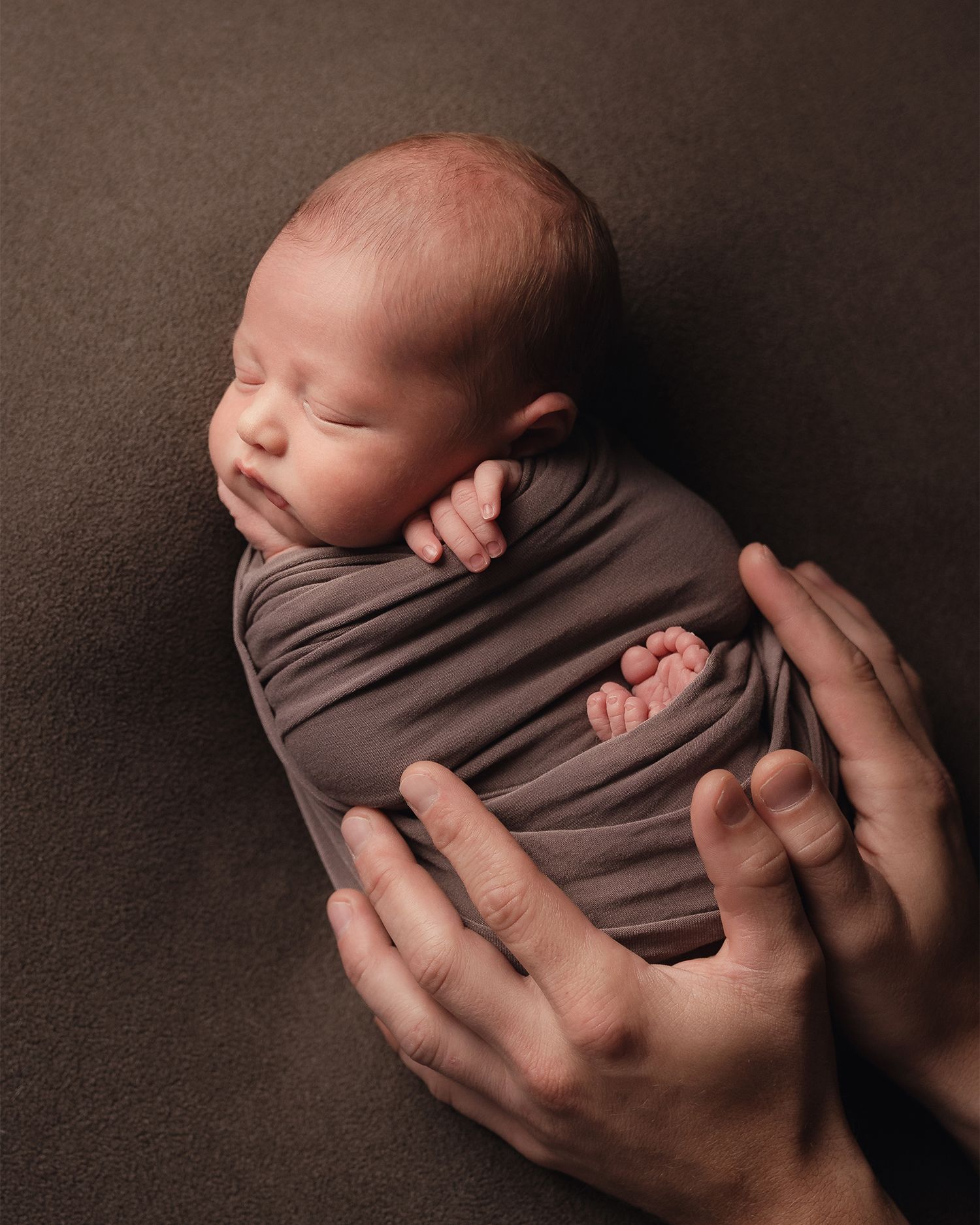 Newborn baby swaddled in gray wrap held gently by parent hands.