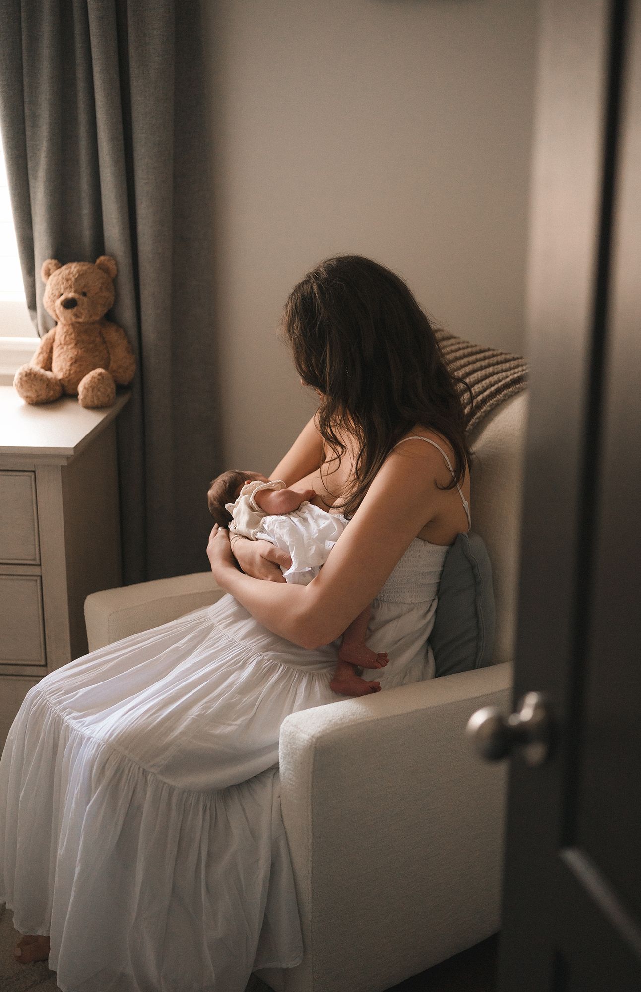 Mother in a white dress sits in a cozy armchair nursing her newborn baby in a softly lit nursery with a teddy bear on the dresser.