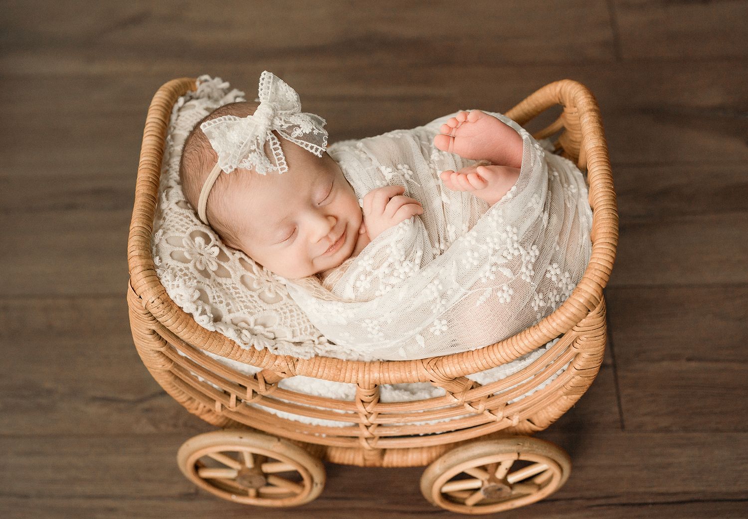 A sleeping newborn girl in lace wrap rests in a wicker carriage prop on a wooden floor.