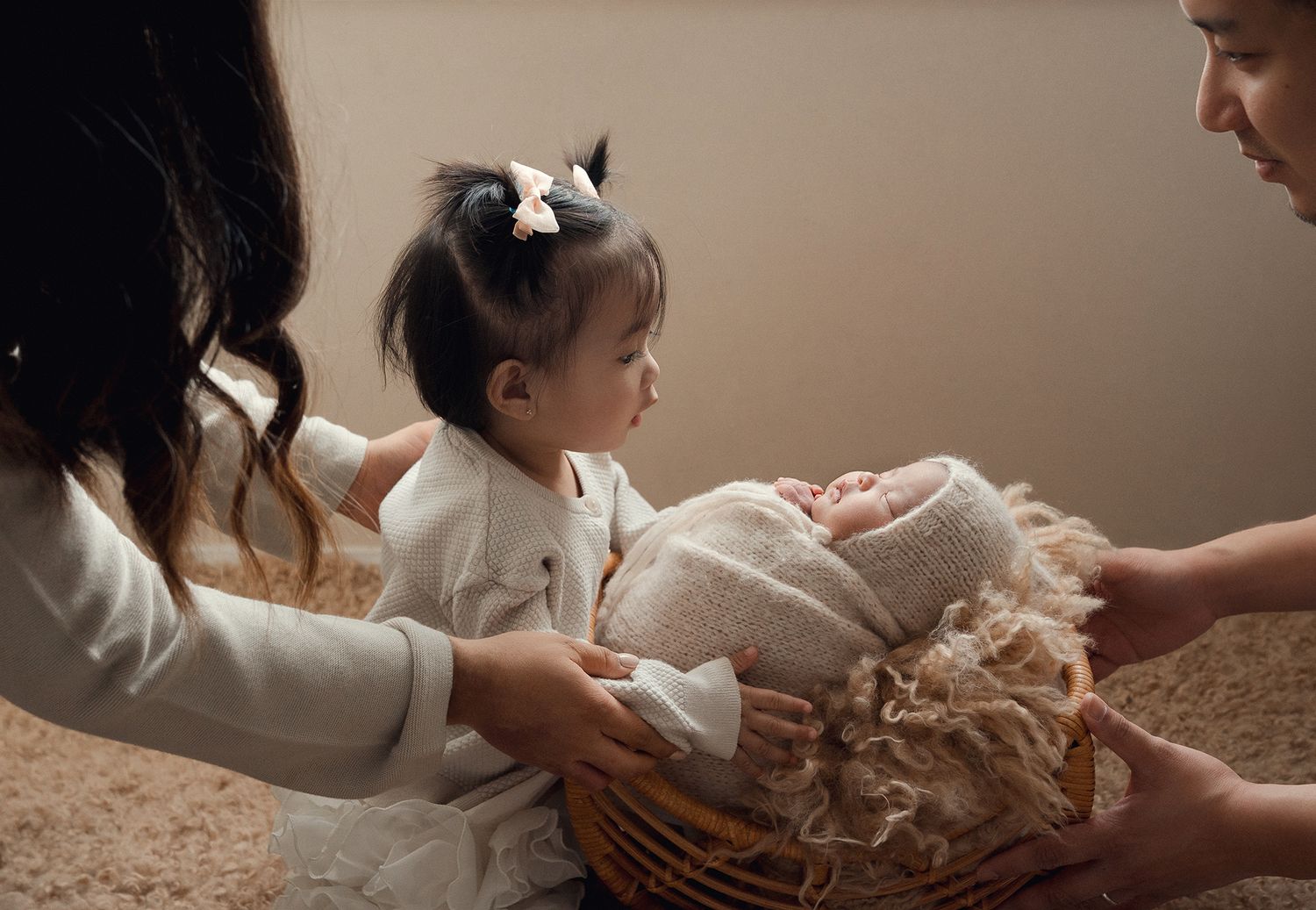 Toddler girl with bow gazes at swaddled newborn sibling held in a basket by parents.