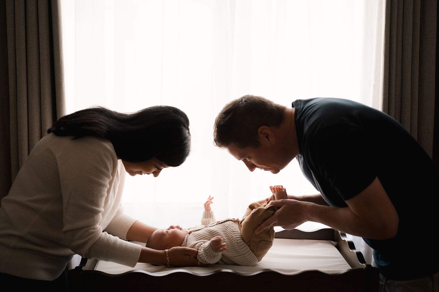 Silhouetted parents lean over a changing table to smile at their baby in soft natural window light.
