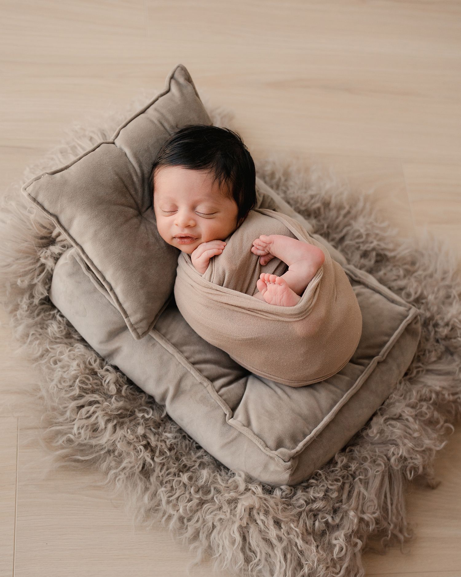 Peaceful newborn baby swaddled in beige wrap, sleeping on a miniature bed with fluffy gray rug in a professional photo.