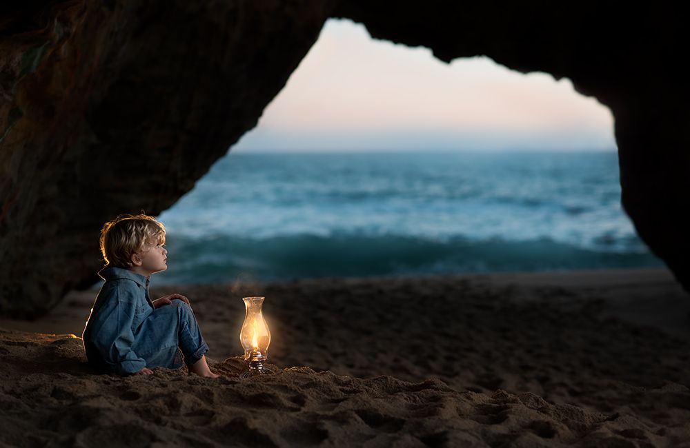 Young boy sitting in sea cave at dusk holding a glowing lantern by ocean.