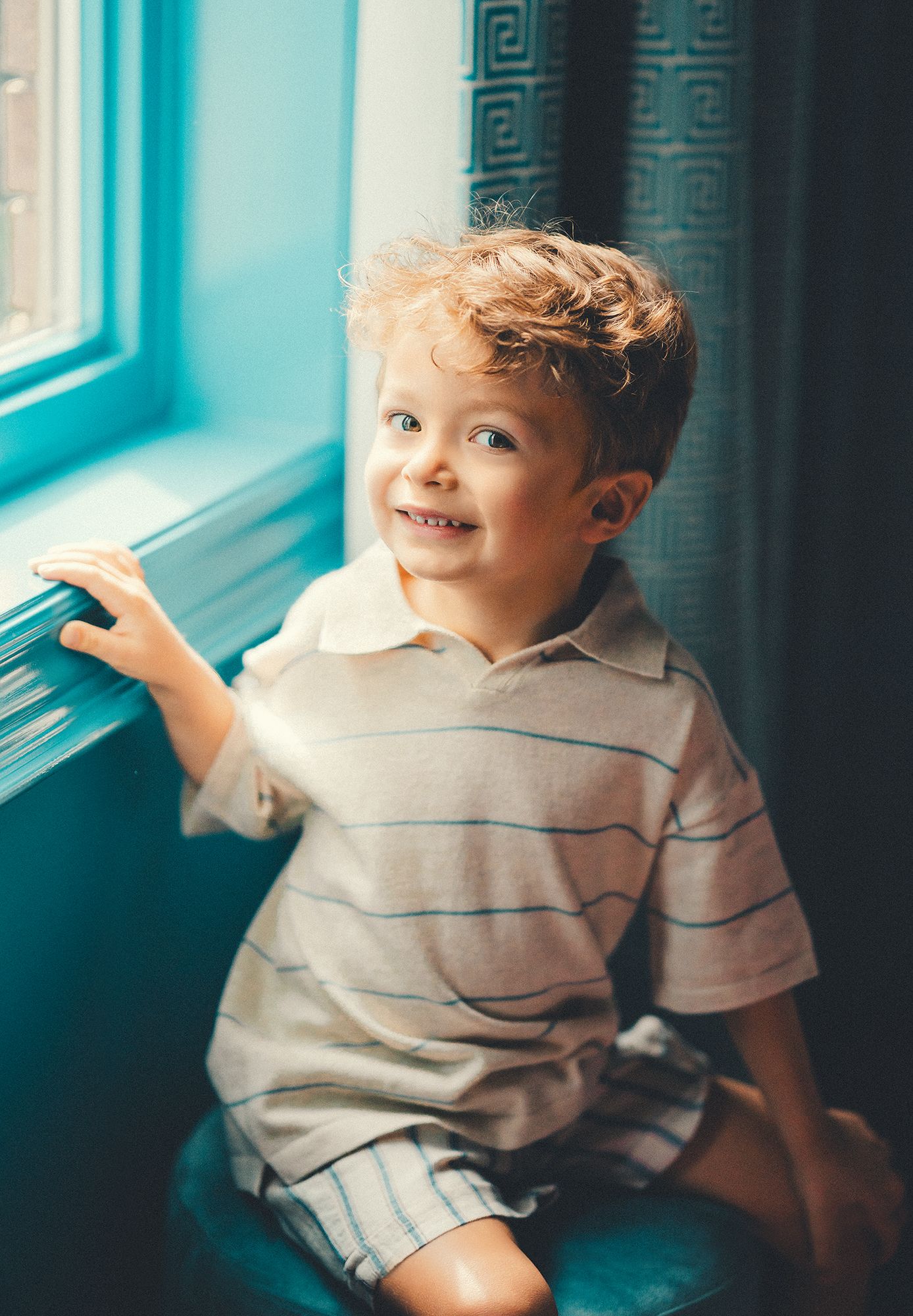 A cheerful curly-haired boy sits by a bright teal window, smiling and touching the sill.
