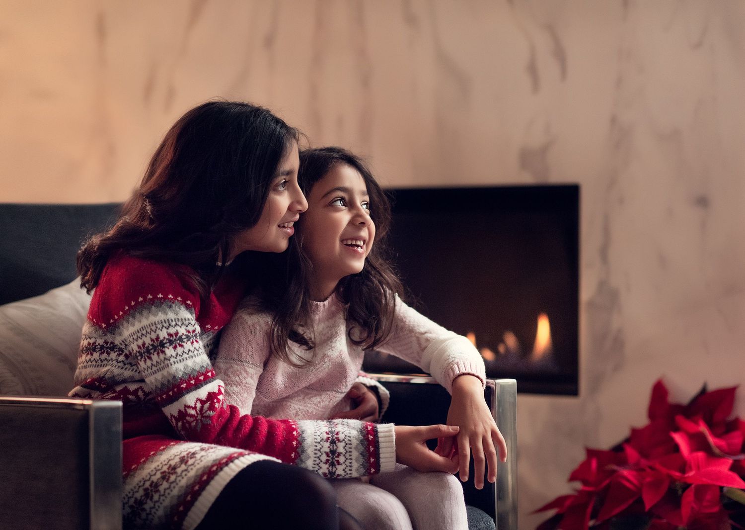 Two young sisters in Christmas sweaters sit together by a cozy fireplace with holiday decorations.