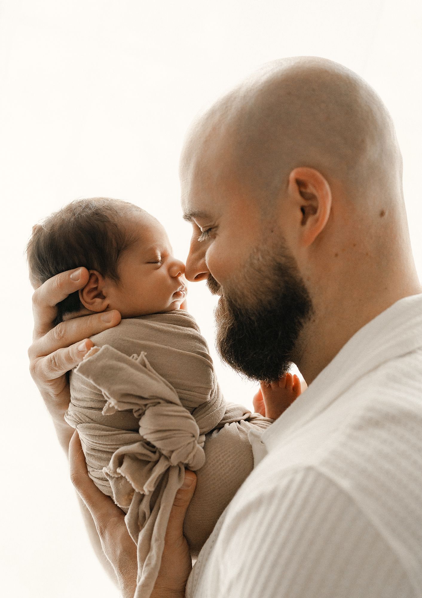 Bearded bald father gently touches noses with his sleeping newborn baby wrapped in a soft beige swaddle against a bright background.