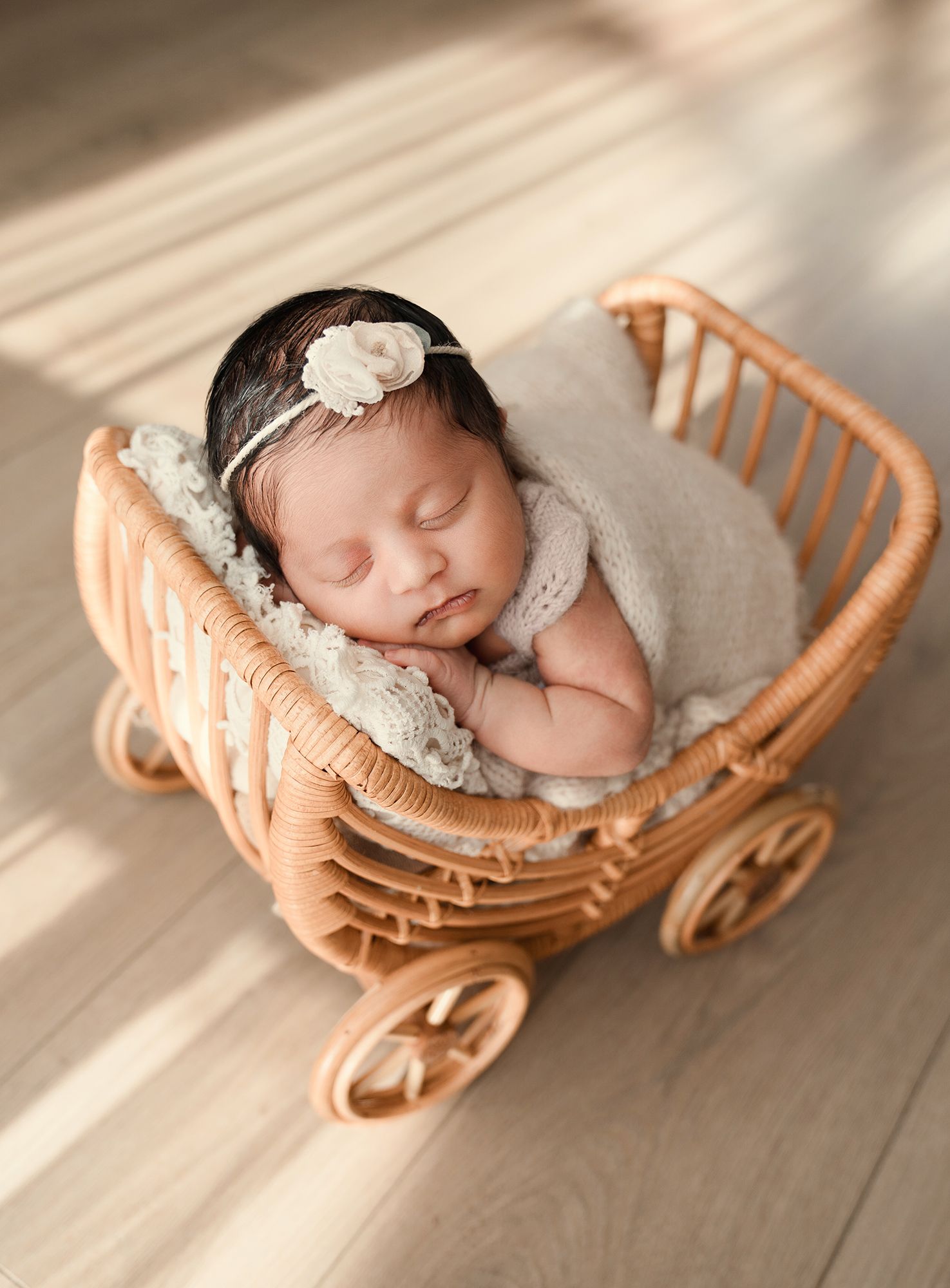 A sleeping newborn baby girl in a cream wrap rests in a miniature wicker pram prop.
