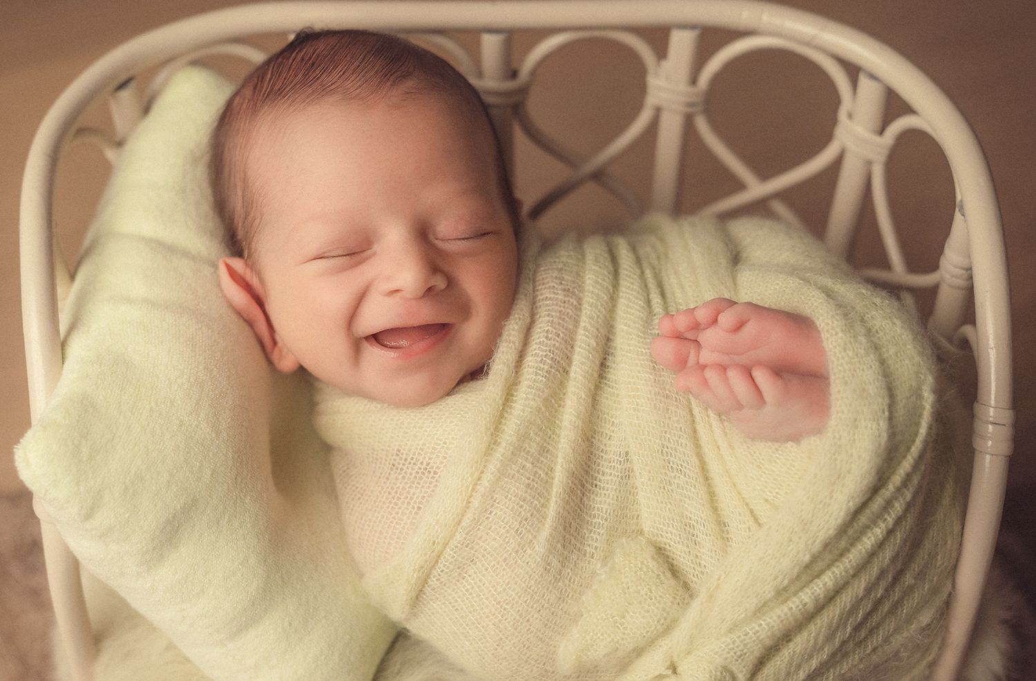 Smiling newborn baby wrapped in light green swaddle blanket in a white chair.