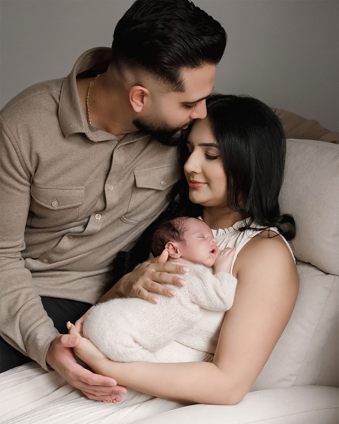 A couple tenderly gazes at their sleeping newborn baby wrapped in white, sitting together on a cream sofa.