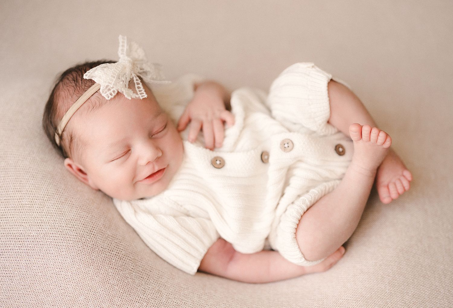 Sleeping newborn baby girl in a cream knit romper and lace bow headband curled up and smiling on a soft neutral backdrop.