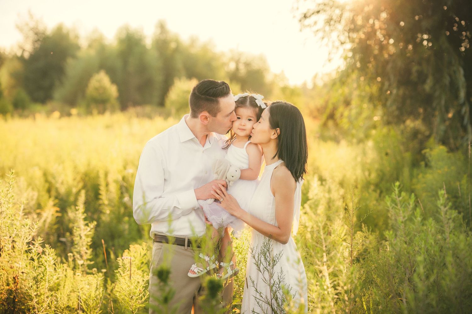 Parents kissing their young daughter in a sunlit green meadow at golden hour.