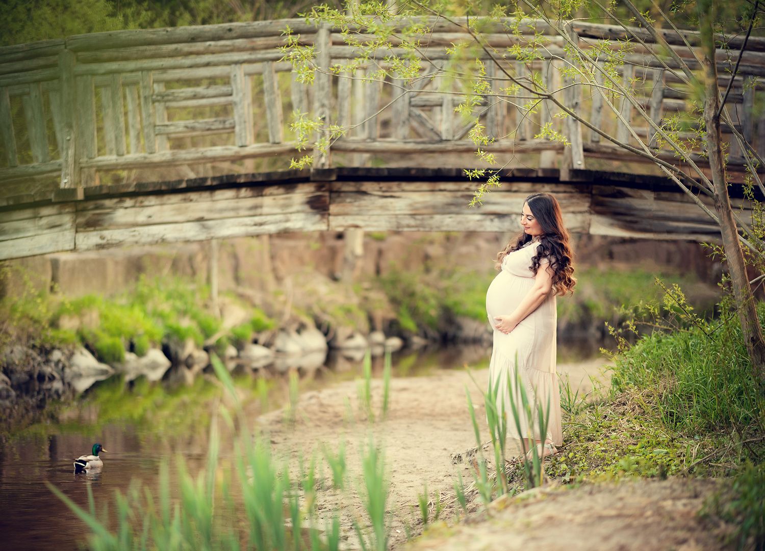 Pregnant woman in white dress standing near rustic bridge by peaceful creek.