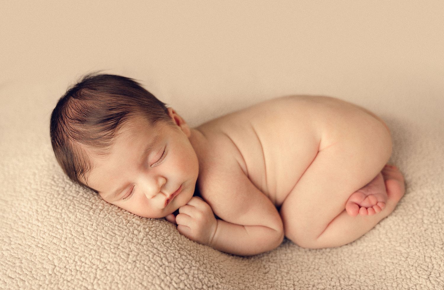 Peaceful naked newborn baby sleeping curled up on a soft beige blanket in a classic pose.