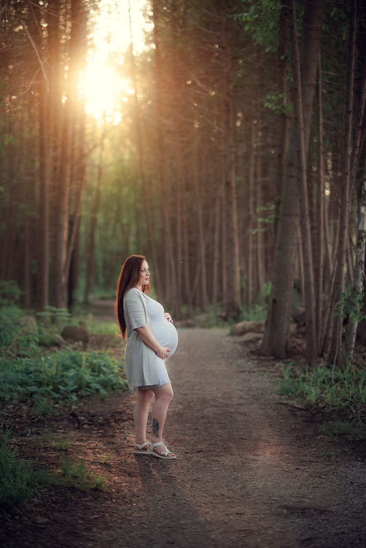 Pregnant woman in white dress standing on forest path bathed in golden sunset light.
