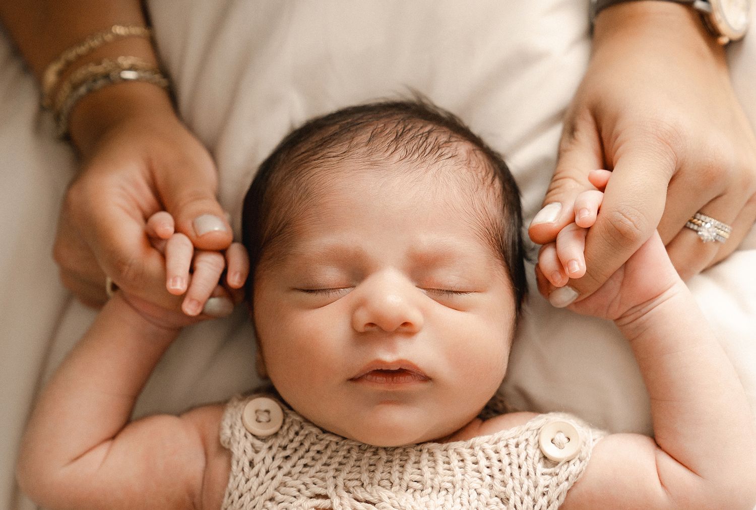 Sleeping newborn baby in a knit romper holding the fingers of two adults' hands against soft white bedding.