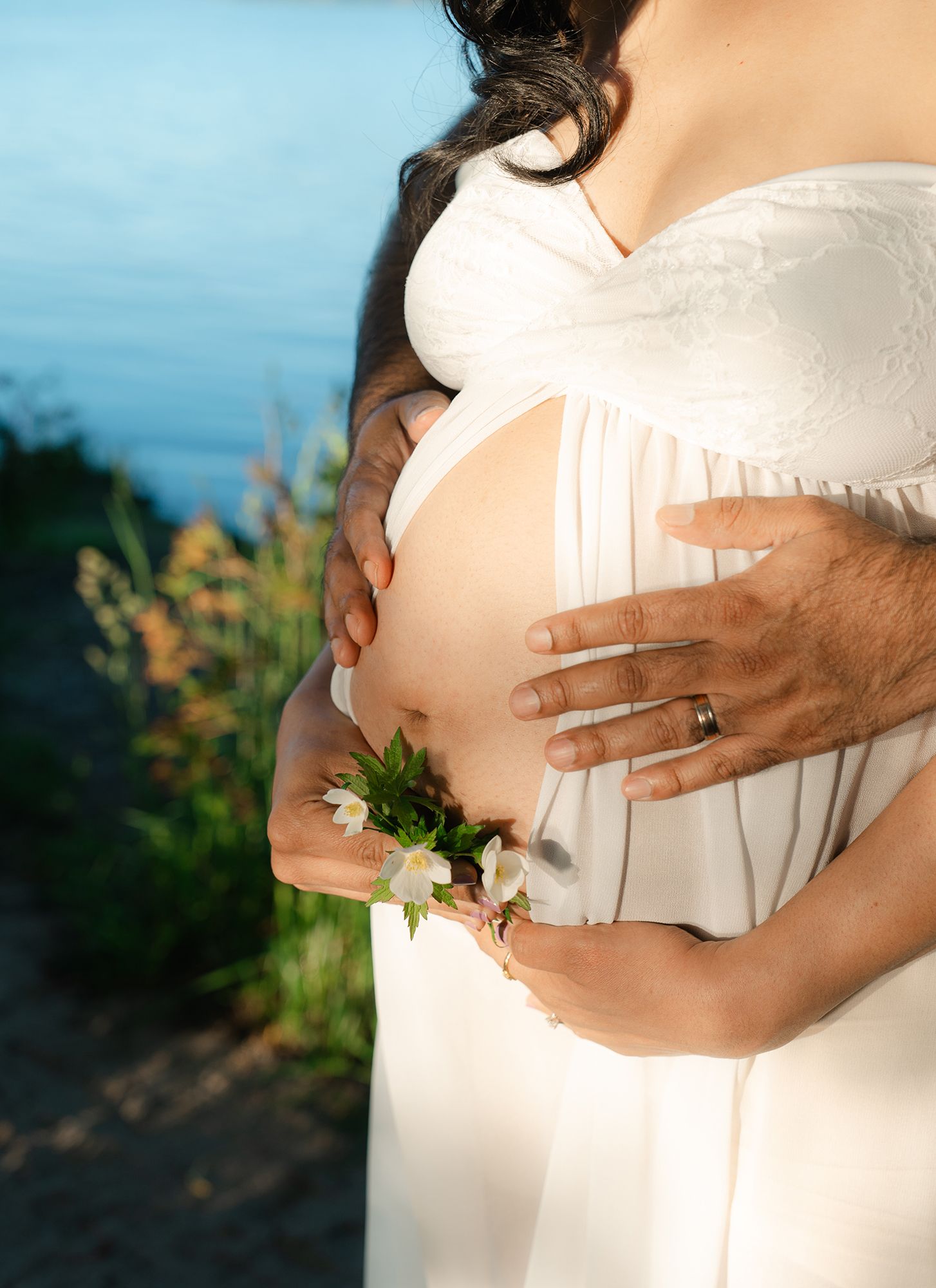 Pregnant woman in white dress with partner's hands on baby bump, adorned with small white flowers, near water.