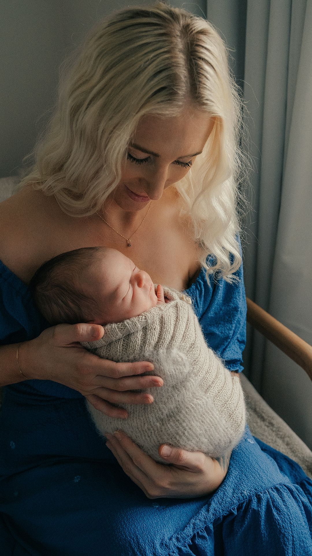 A blonde mother in a blue dress tenderly holds her sleeping newborn baby wrapped in a cozy knit blanket.