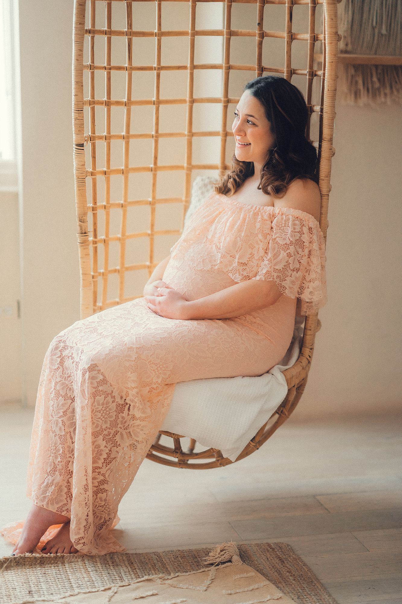 Smiling pregnant woman in pink lace dress sitting in rattan hanging chair.