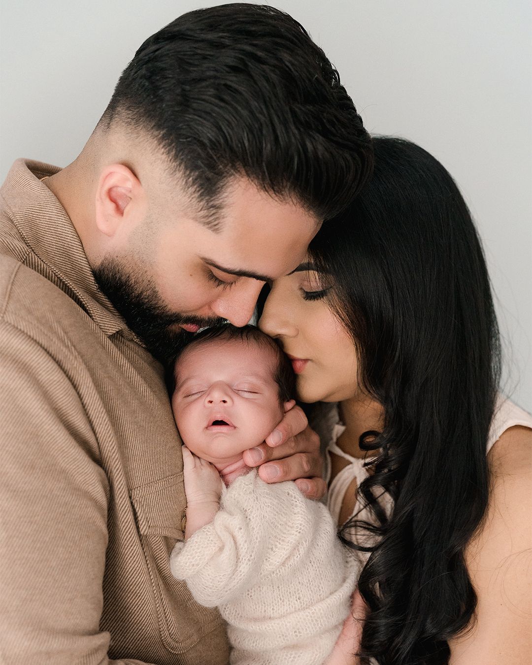 New parents in neutral tones lovingly embrace their sleeping newborn baby together.