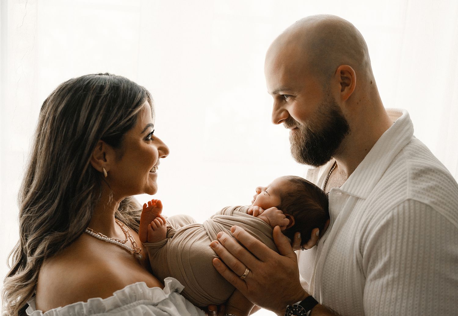 Smiling couple in white outfits lovingly holds their newborn baby in soft natural window light.