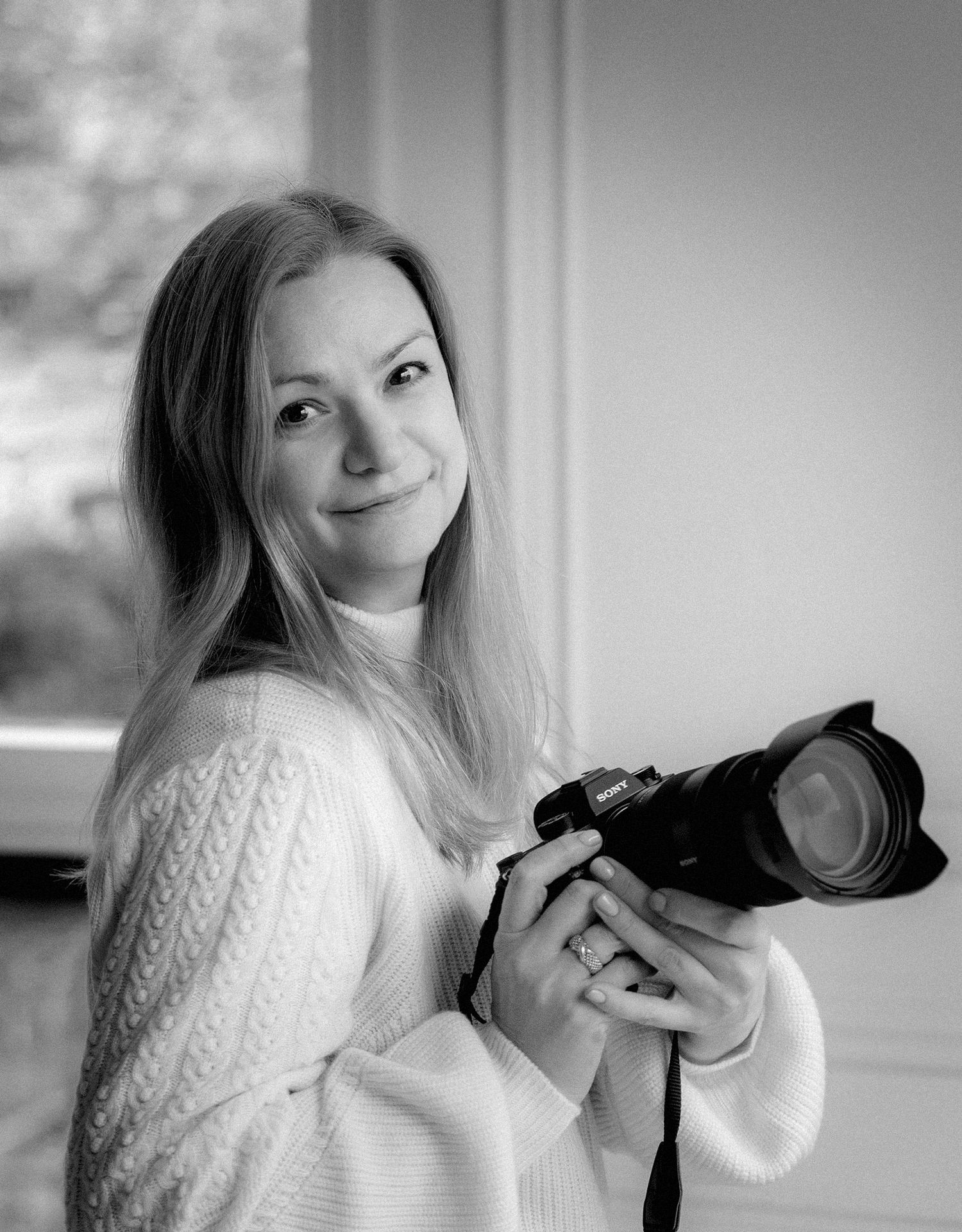 Black and white portrait of blonde female photographer smiling while holding Sony camera indoors.