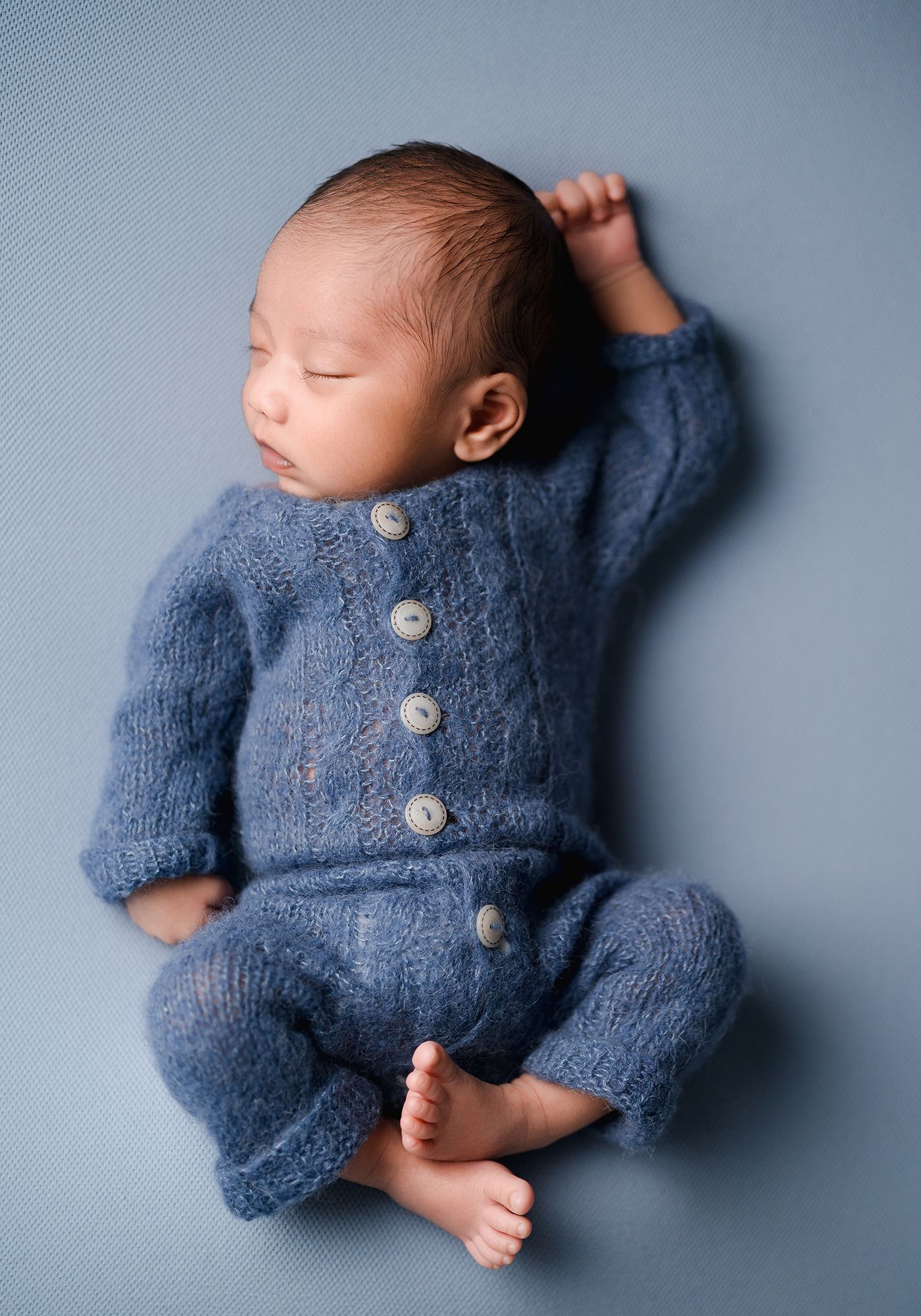 Sleeping newborn baby in a blue knitted button-up romper lying on a soft blue backdrop during a newborn photo session.